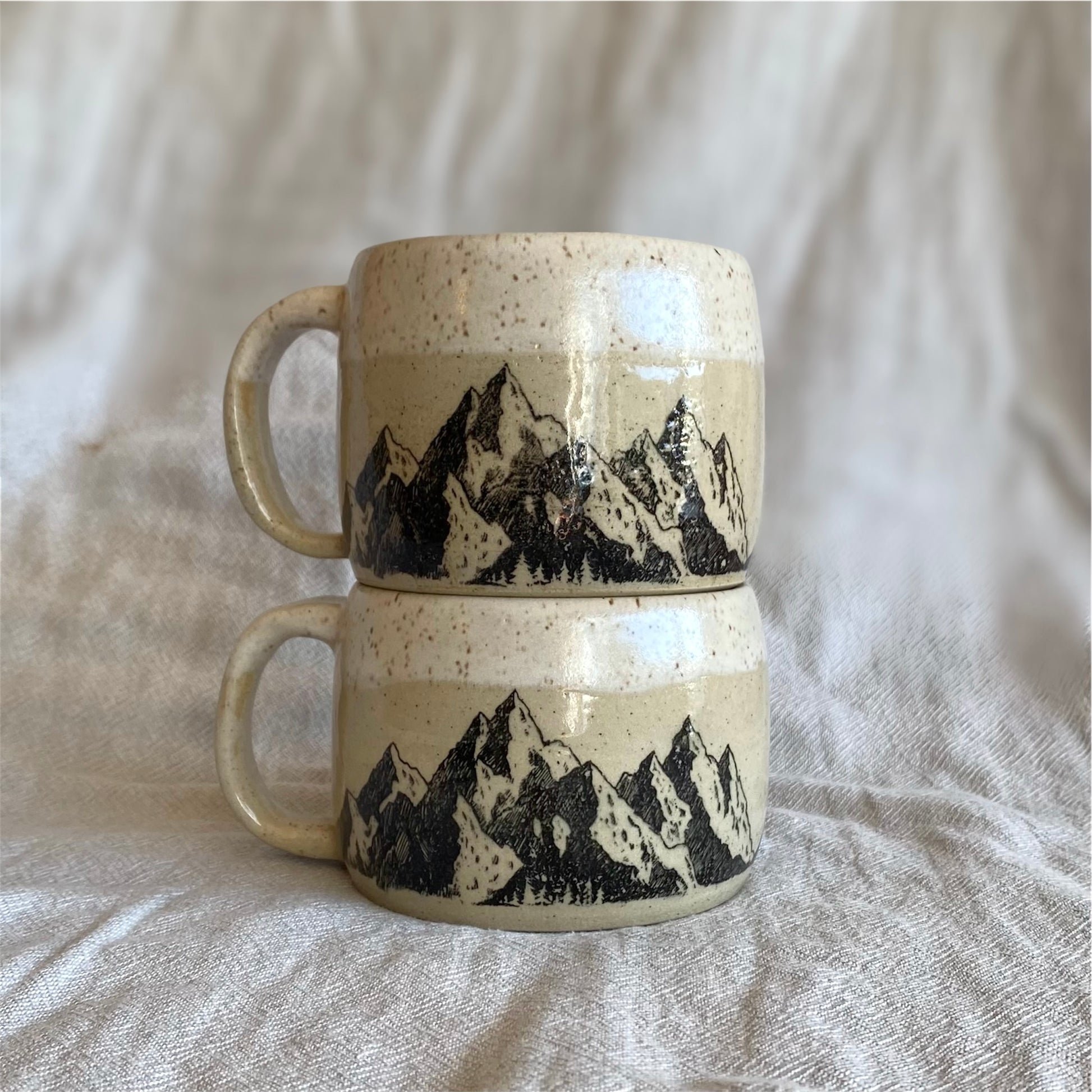 Two ceramic mugs with mountain design stacked on a textured fabric background