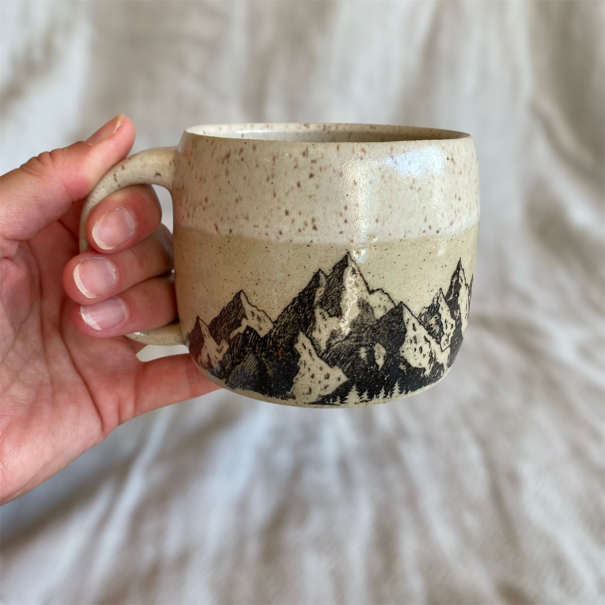 Hand holding a ceramic mug with mountain design against a neutral background