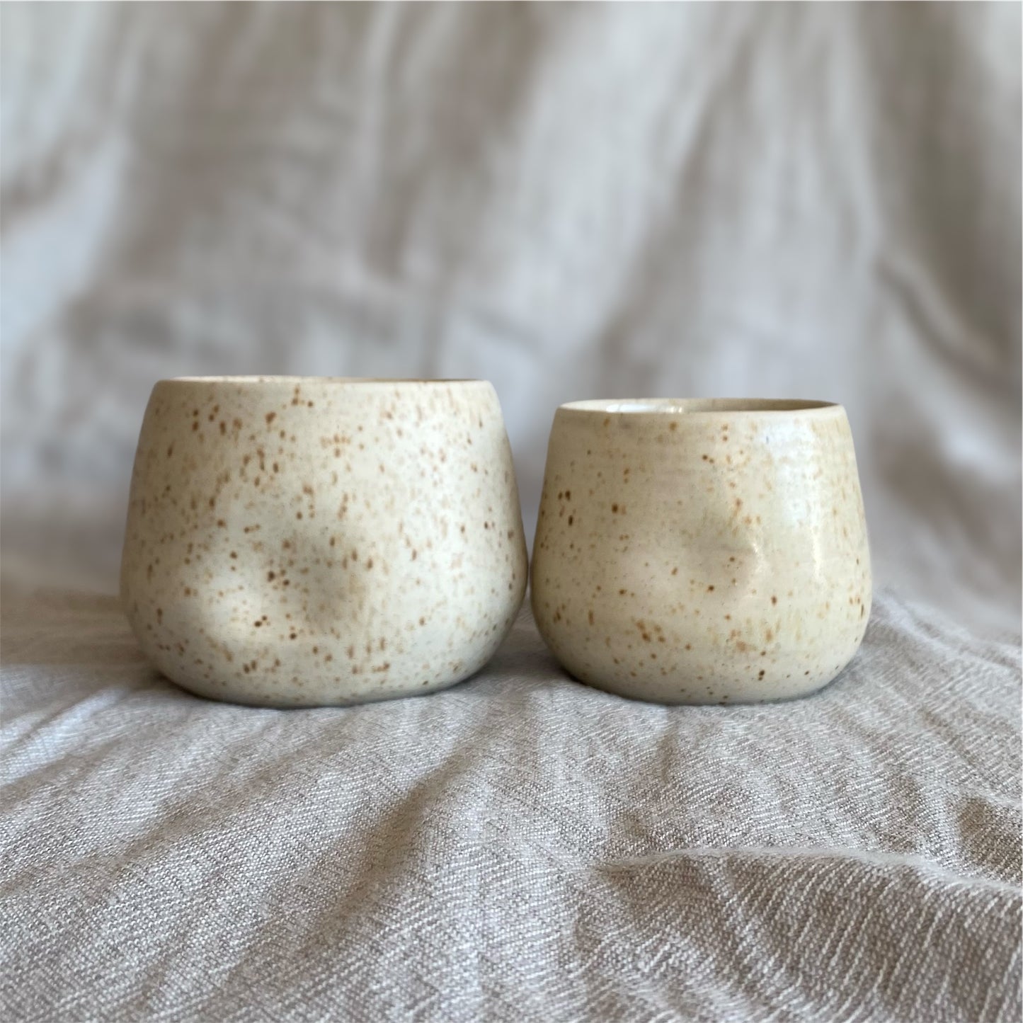 Two ceramic cups with a speckled texture on a textured fabric background