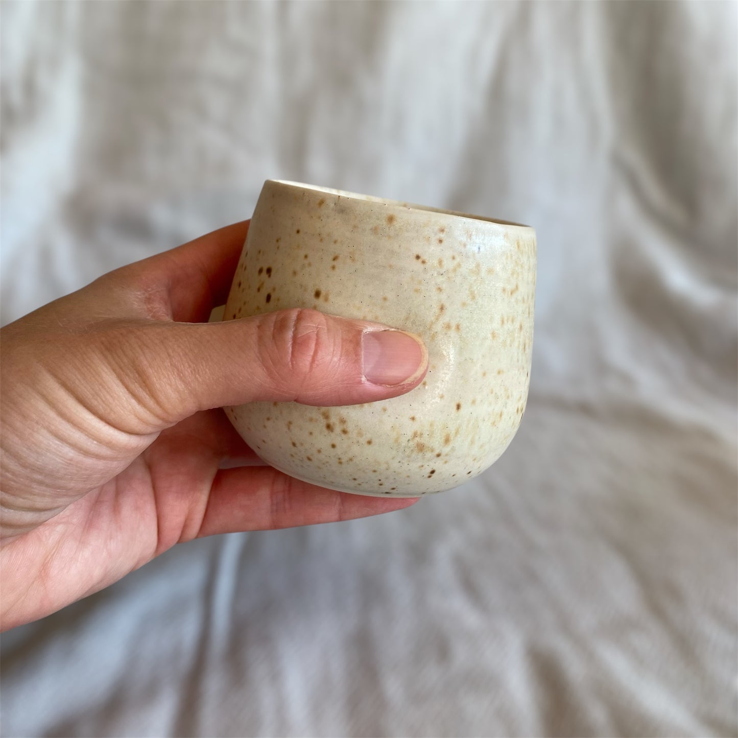 Hand holding a small ceramic cup with a speckled texture against a neutral background