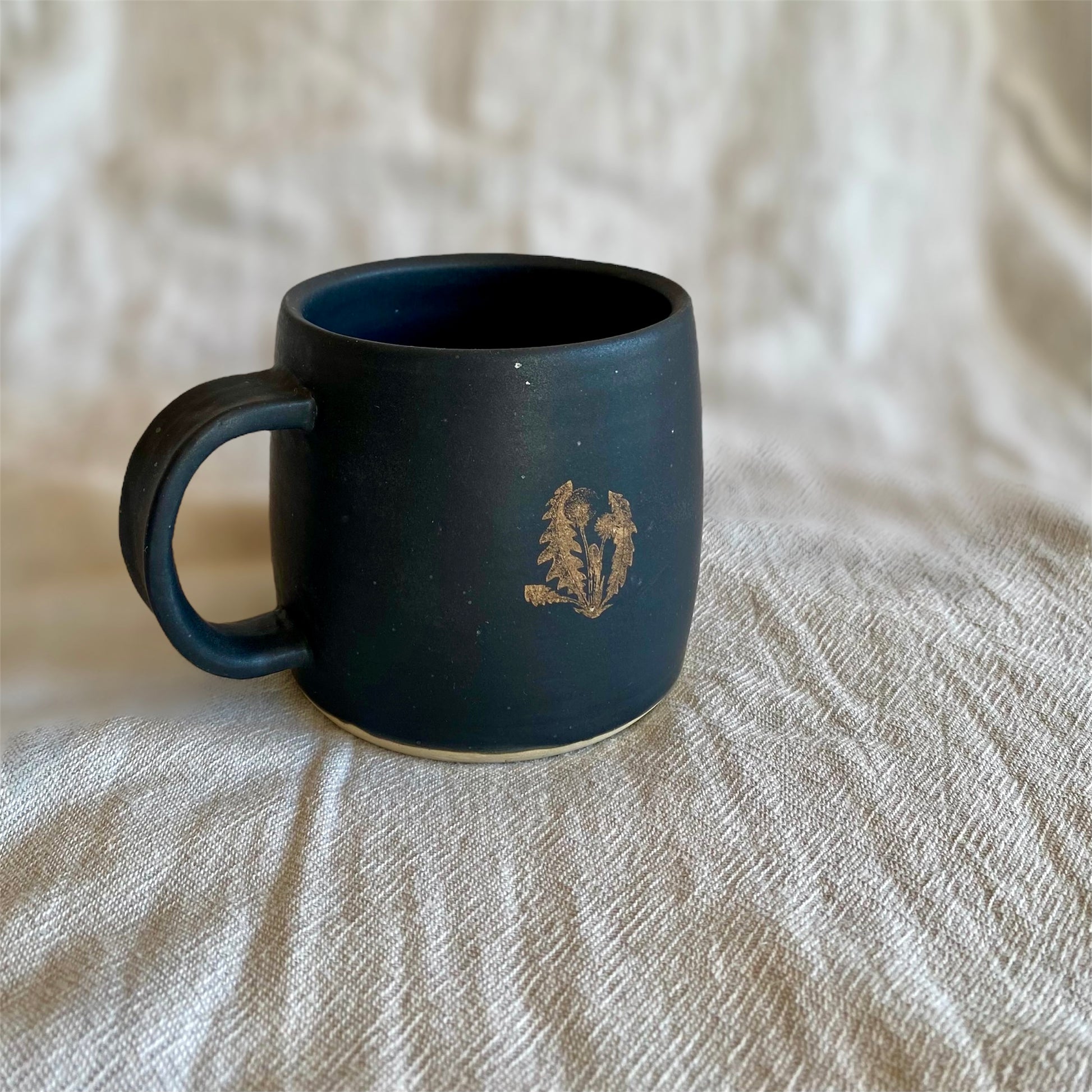 Black mug with a gold dandelion on a neutral textured surface