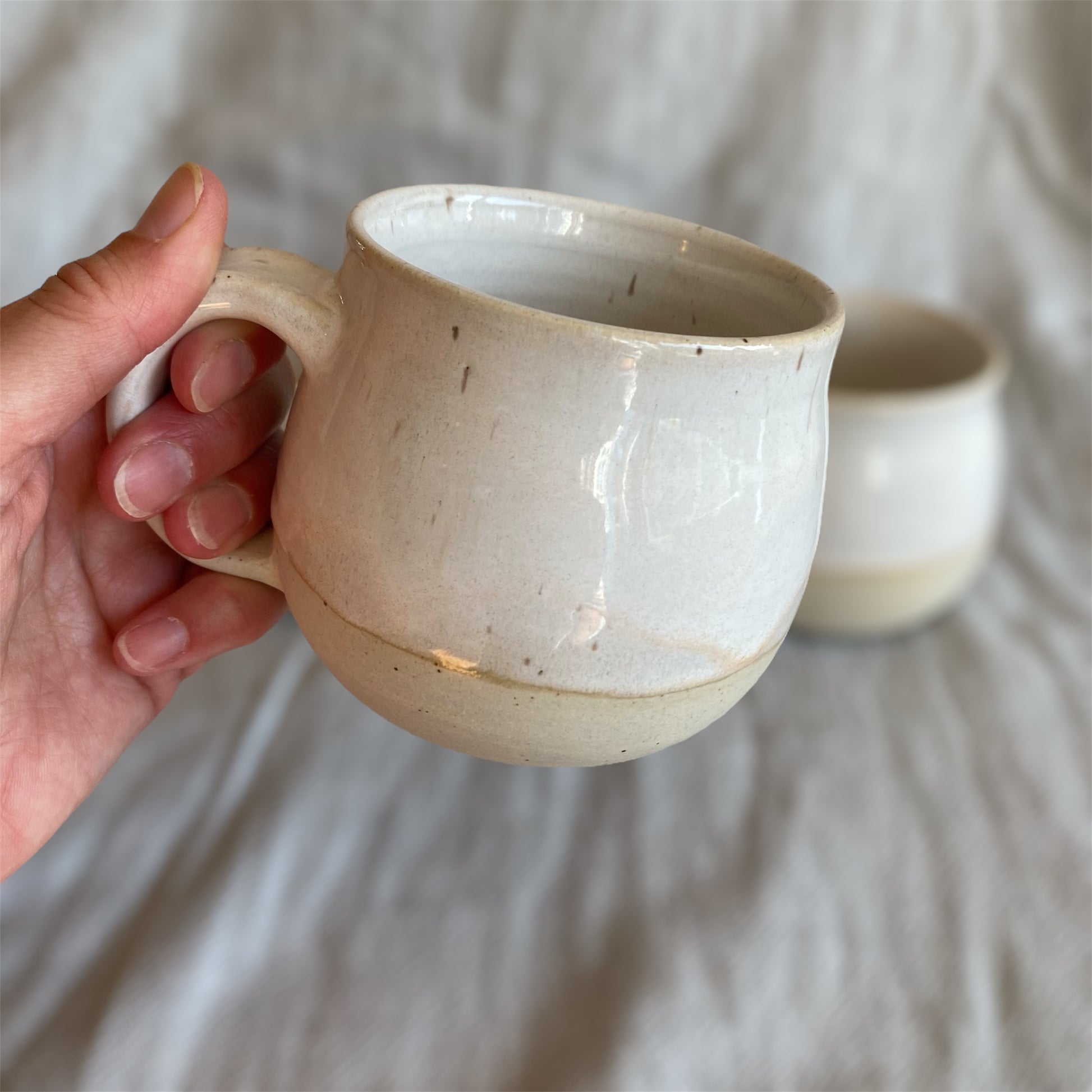 Hand holding a ceramic mug with another mug in the background on a neutral fabric surface
