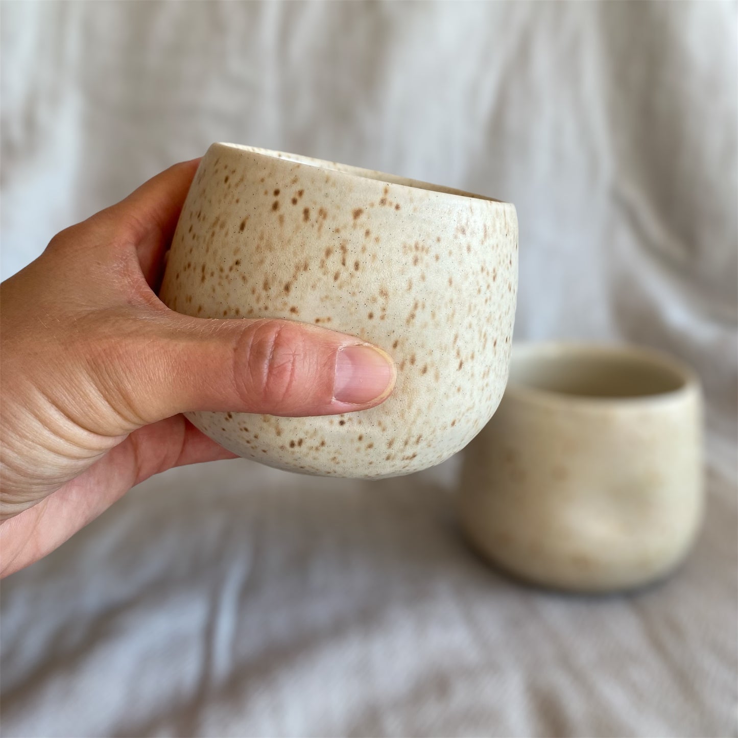 Hand holding a textured ceramic cup with another cup in the background on a neutral surface