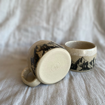 Two ceramic mugs with mountain designs on a textured surface