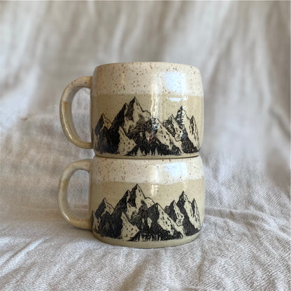 Two ceramic mugs with mountain design stacked on a textured fabric background