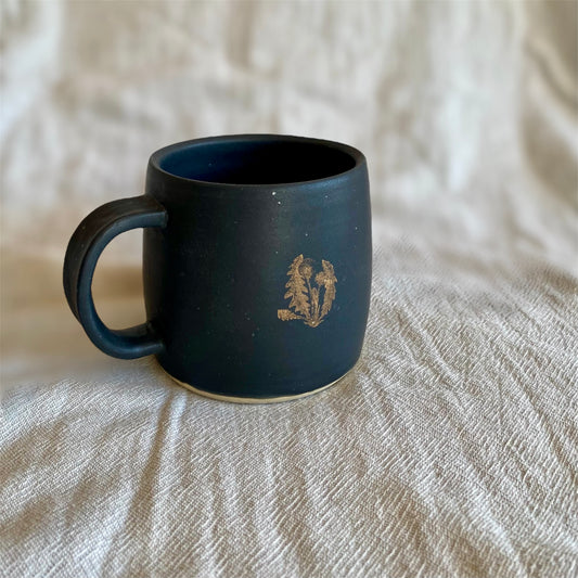 Black mug with a gold dandelion on a neutral textured surface