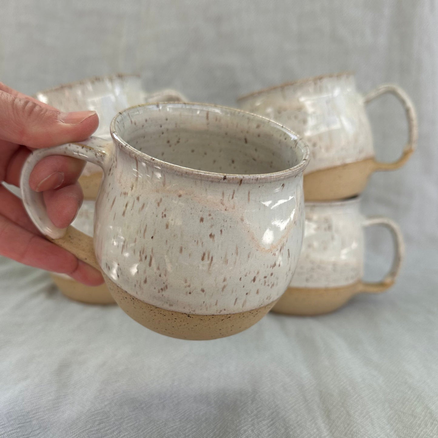 Hand holding a ceramic mug with speckled texture on a light fabric background